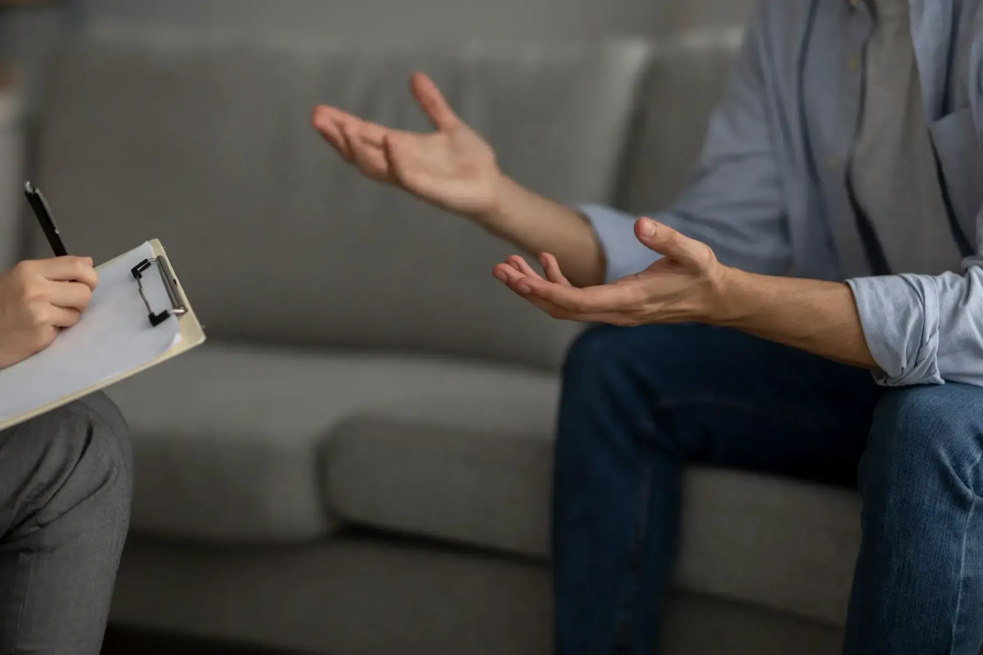 Una persona habla, gesticula con las manos, mientras otra toma notas en un portapapeles; ambos están sentados en un sofá.