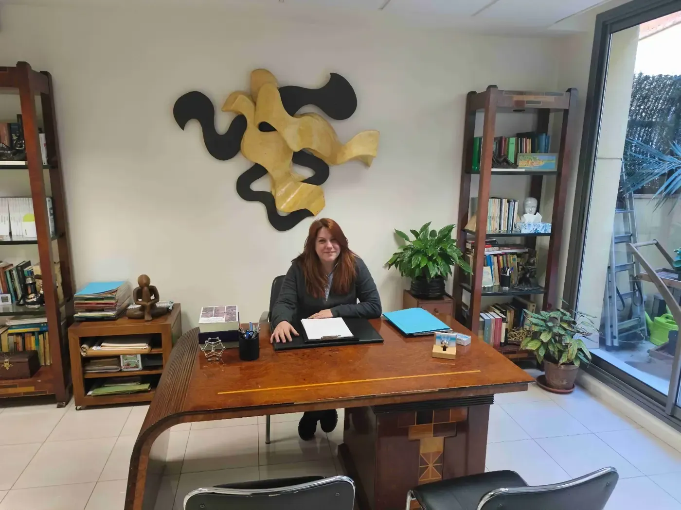 Mujer sentada en un escritorio de madera en una oficina. Se ven estanterías, arte mural abstracto y una planta.