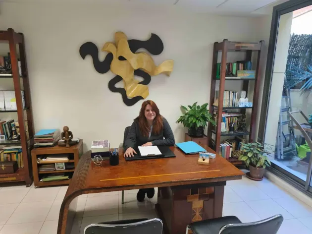 Mujer sentada en un escritorio de madera en una oficina. Se ven estanterías, arte mural abstracto y una planta.