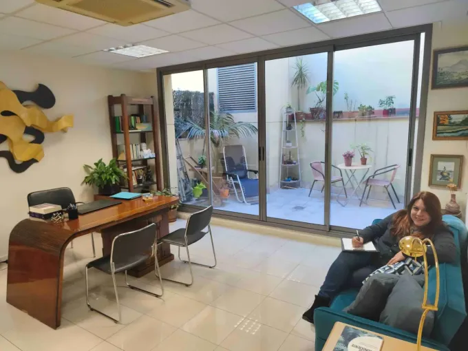 Mujer escribiendo en un sofá en una oficina con un escritorio, estantes y vista a un pequeño patio al aire libre.