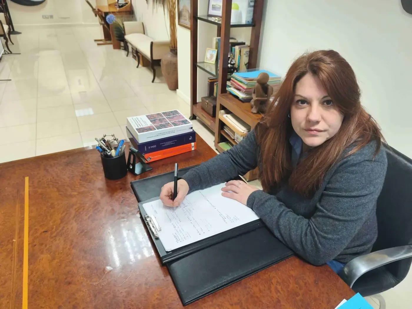 Mujer escribiendo en un escritorio, mirando a la cámara. Oficina con libros y estanterías.