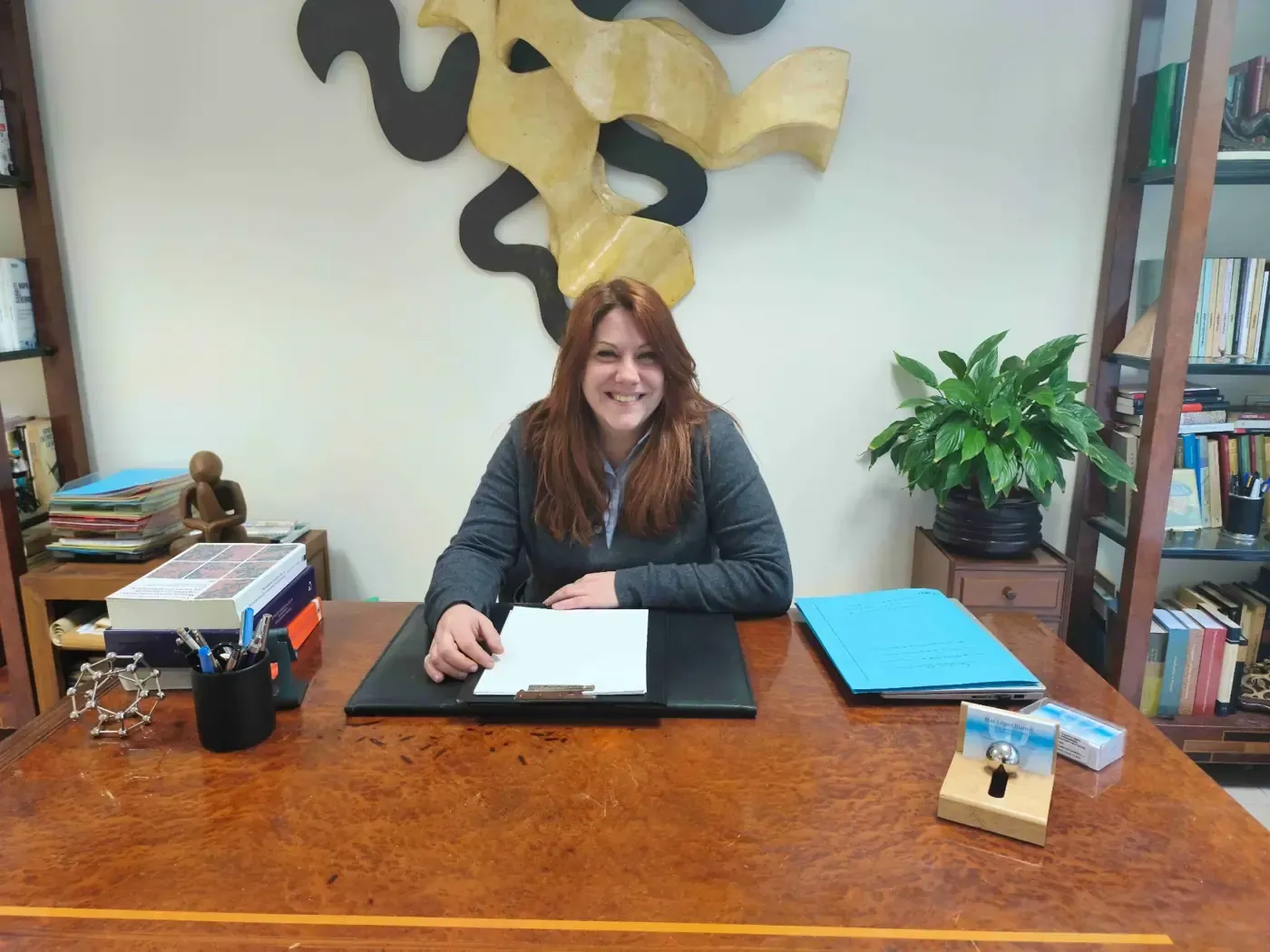 Mujer sentada en un escritorio de madera, sonriendo. Obras de arte y plantas de fondo.