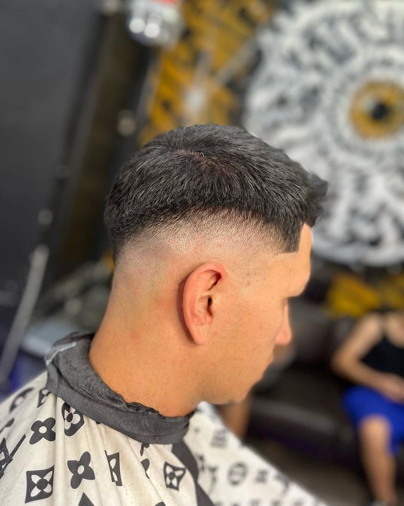 Hombre con un corte de pelo descolorido, vestido con una capa de barbero, sentado en una barbería.