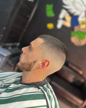 Hombre con corte de pelo descolorido, barba y pendiente, con capa de barbero. Fondo de barbería con arte mural.