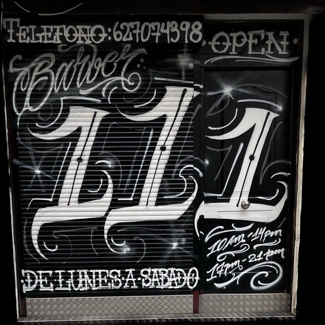 Puerta de barbería en blanco y negro con letras estilizadas y horario. Se ve la palabra "Barber".