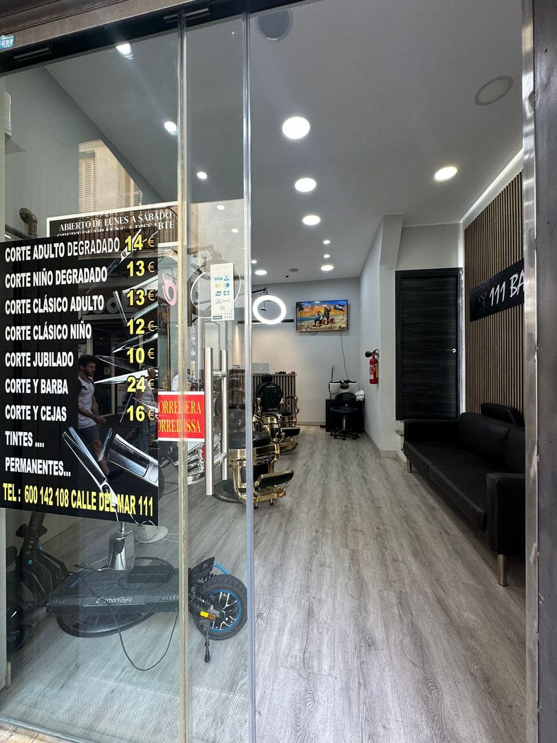 Interior de una barbería visible a través de puertas de vidrio; lista de precios a la izquierda, sofá negro y silla de barbero en el interior.