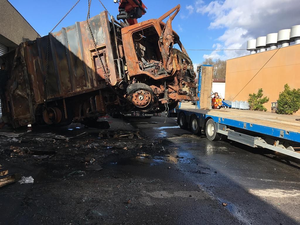 Camion incendié chargé sur une remorque à plateau par une grue ; débris noirs au sol ; journée ensoleillée.