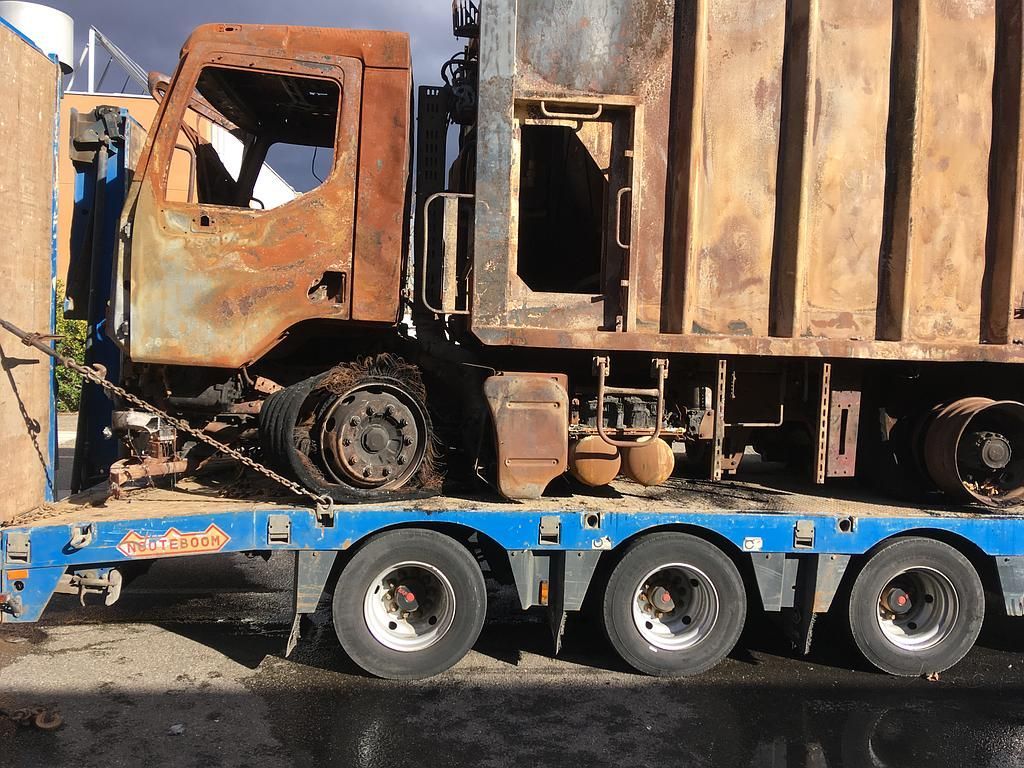 Camion-poubelle incendié sur une remorque à plateau ; rouille et dommages visibles.