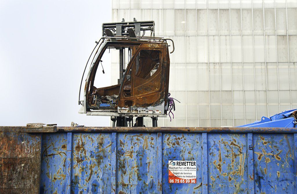 Cabine de grue calcinée sur une benne à ordures bleue, avec un mur en béton en arrière-plan.