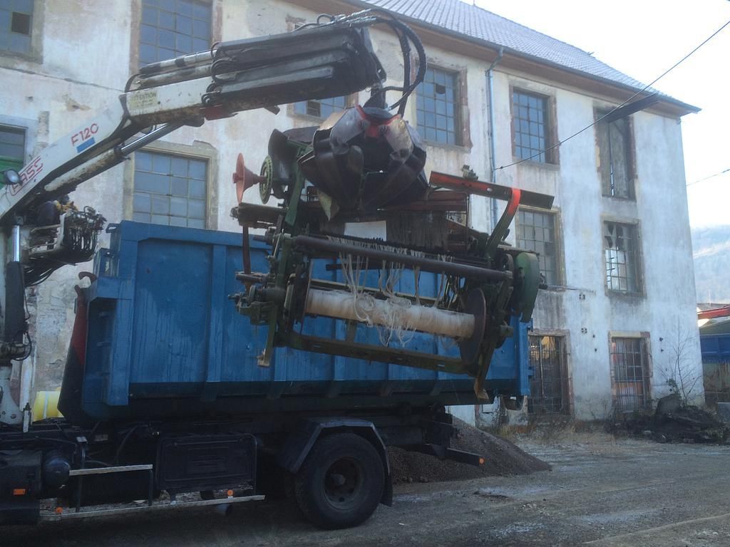 Un camion bleu équipé d'une grue soulève des machines devant un bâtiment délabré.