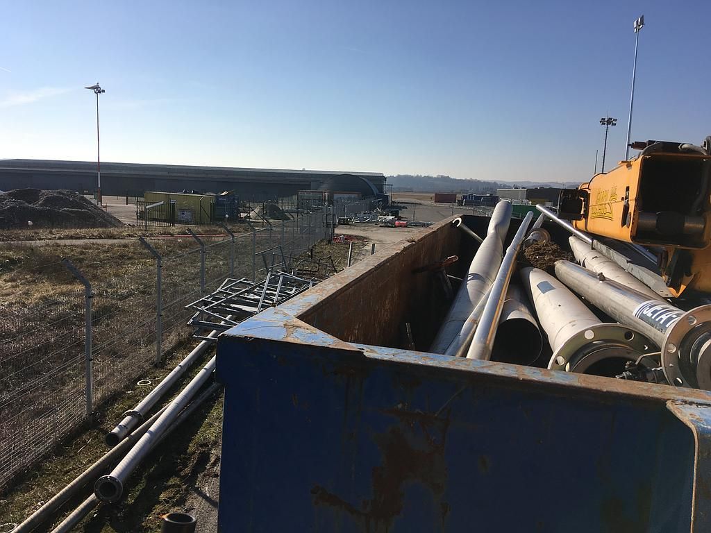Des tuyaux métalliques dans une benne à ordures bleue sur un site industriel par une journée ensoleillée.