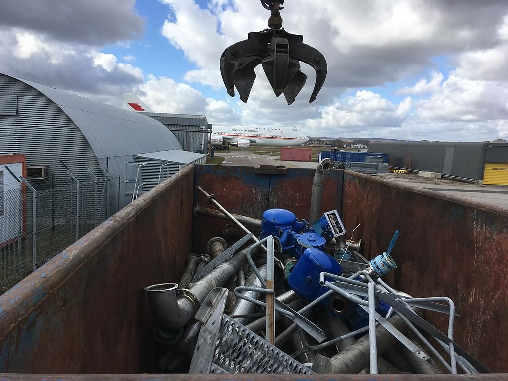 Une pince métallique soulève des déchets métalliques d'un conteneur, l'aéroport étant visible en arrière-plan.