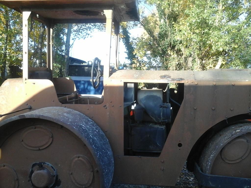 Rouleau compresseur marron rouillé, moteur apparent, stationné à l'extérieur avec des arbres en arrière-plan.