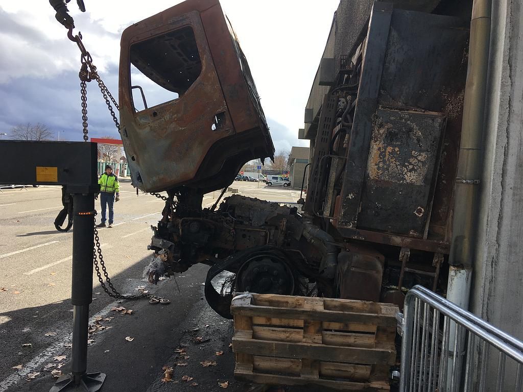 Cabine de camion calcinée soulevée par une grue. Vue extérieure : métal carbonisé, un passant en gilet jaune à proximité.