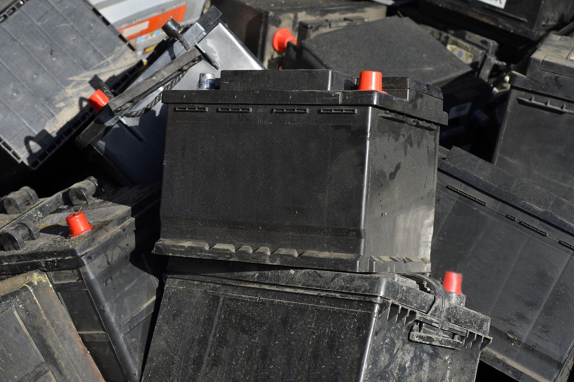 Un tas de batteries de voiture noires, certaines avec des bornes rouges, en extérieur.