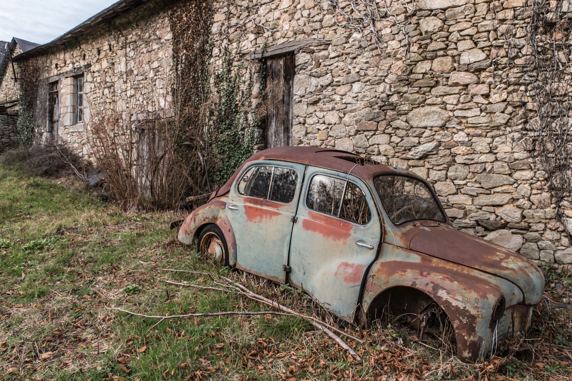 Voiture rouillée abandonnée dans les herbes hautes, à côté d'un bâtiment en pierre.