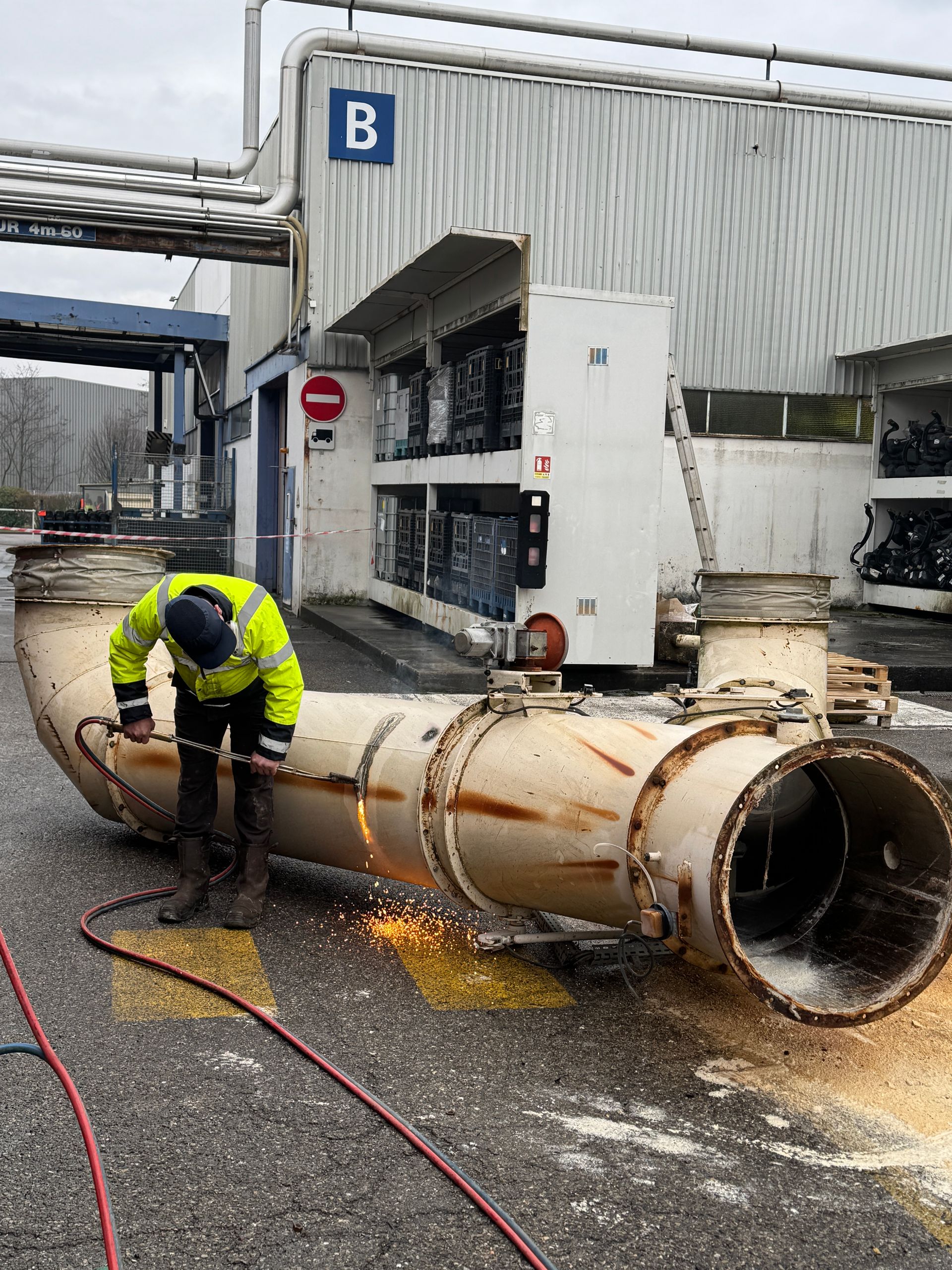 Un ouvrier en tenue de sécurité découpe un gros tuyau métallique à l'extérieur, des étincelles jaillissent ; un bâtiment industriel se trouve en arrière-plan.