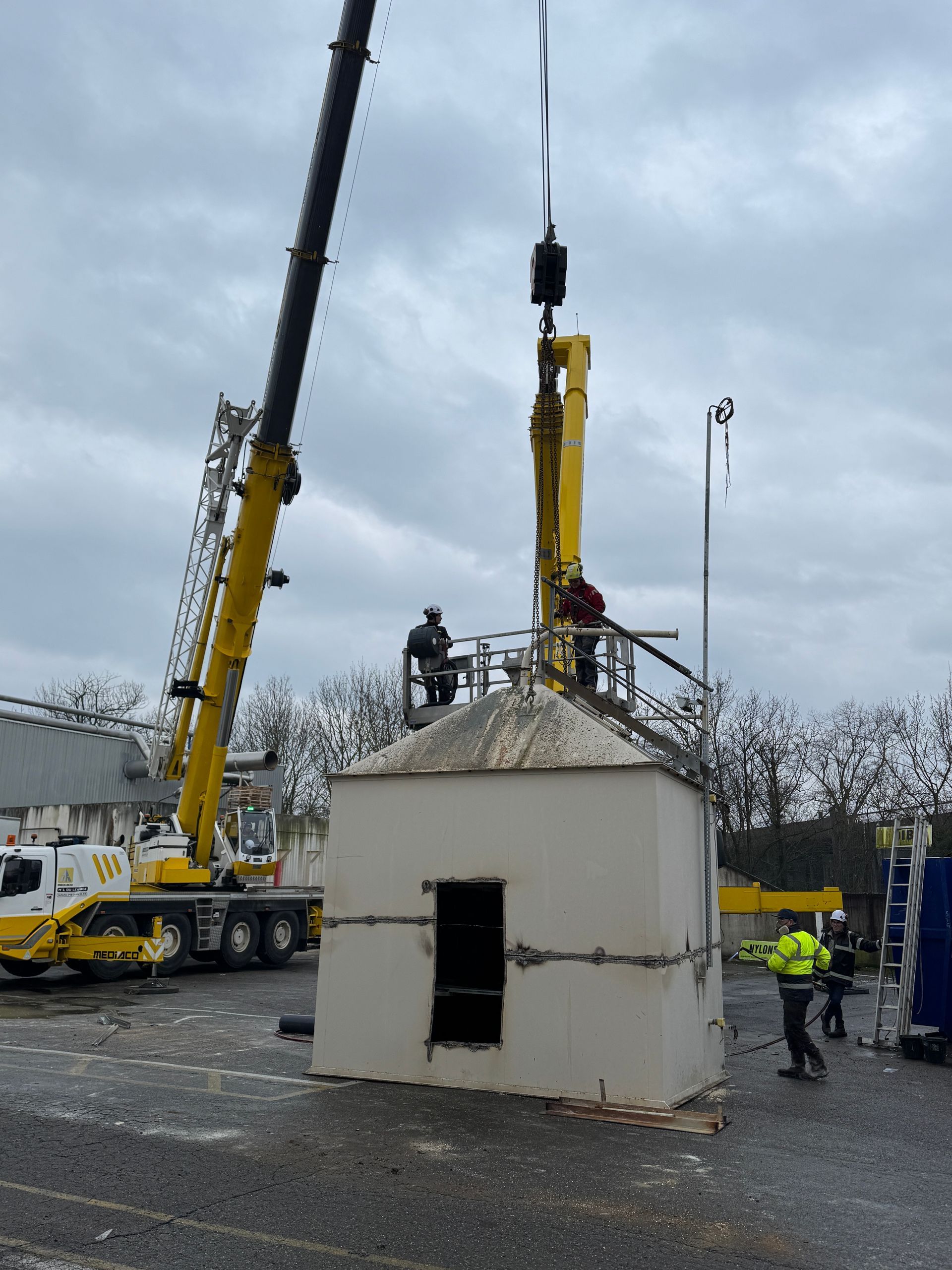 Une grue soulève une structure de silo jaune et blanche ; des ouvriers se trouvent sur la plateforme supérieure et au niveau du sol.