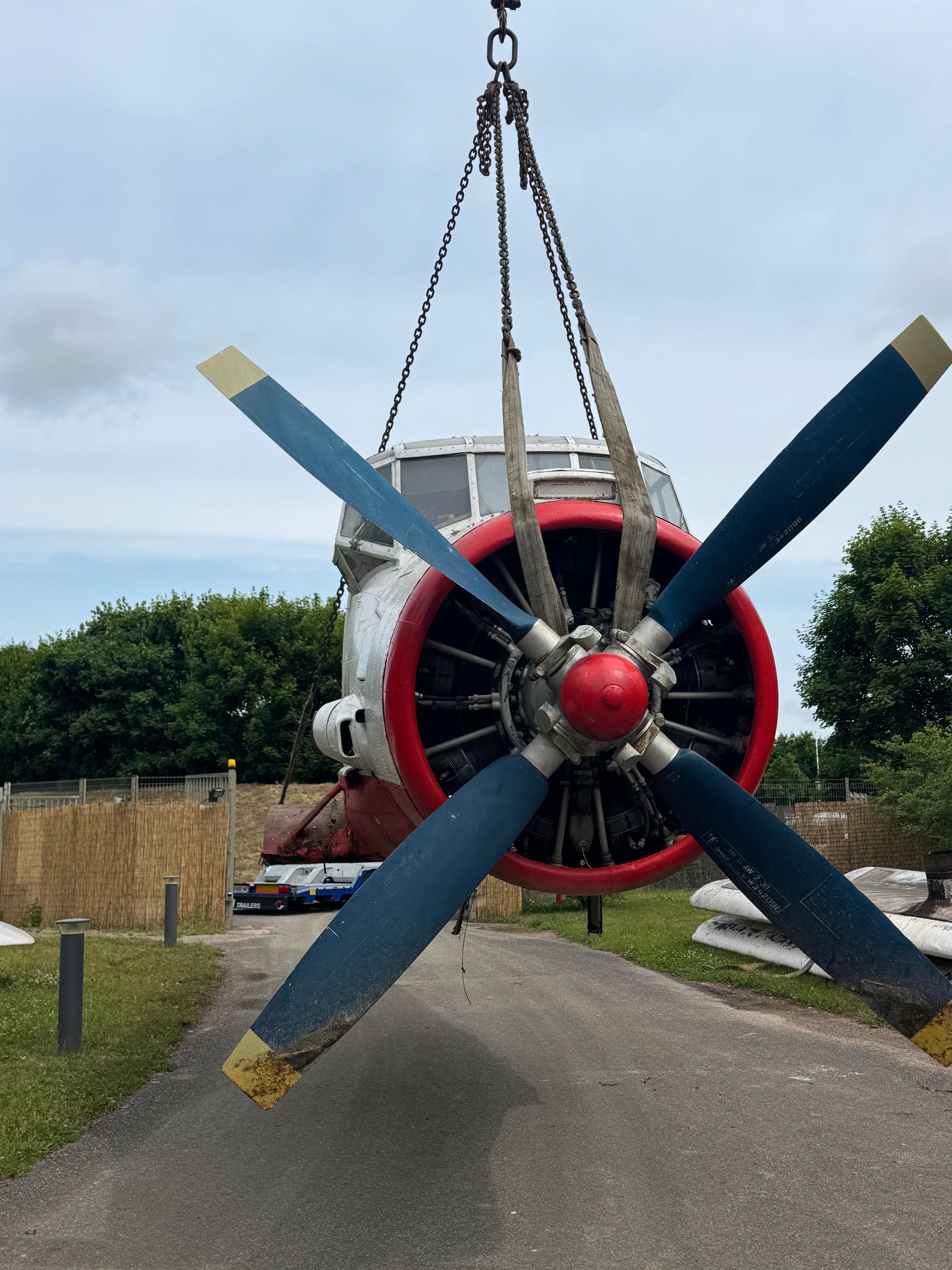 Fuselage d'avion suspendu par des chaînes, pales d'hélice rouges et bleues, décor extérieur.