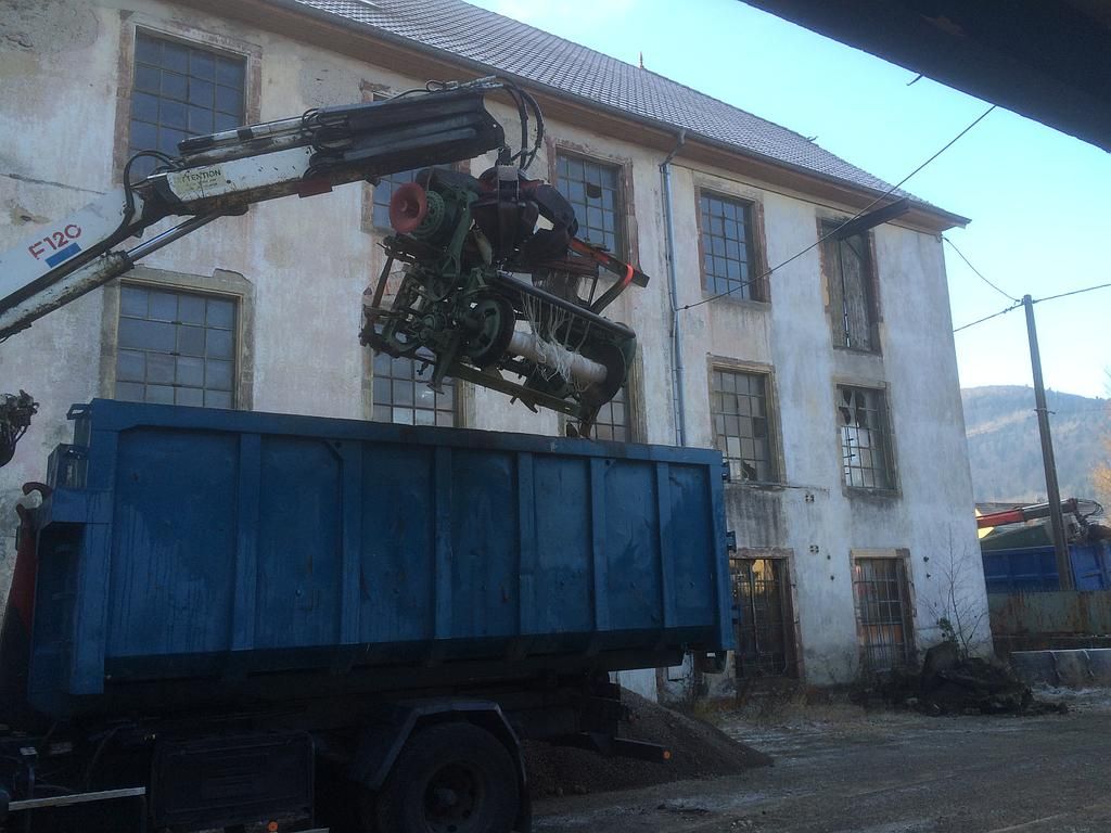 Une grue soulève un moteur et le dépose dans la benne d'un camion bleu devant un vieux bâtiment blanc.