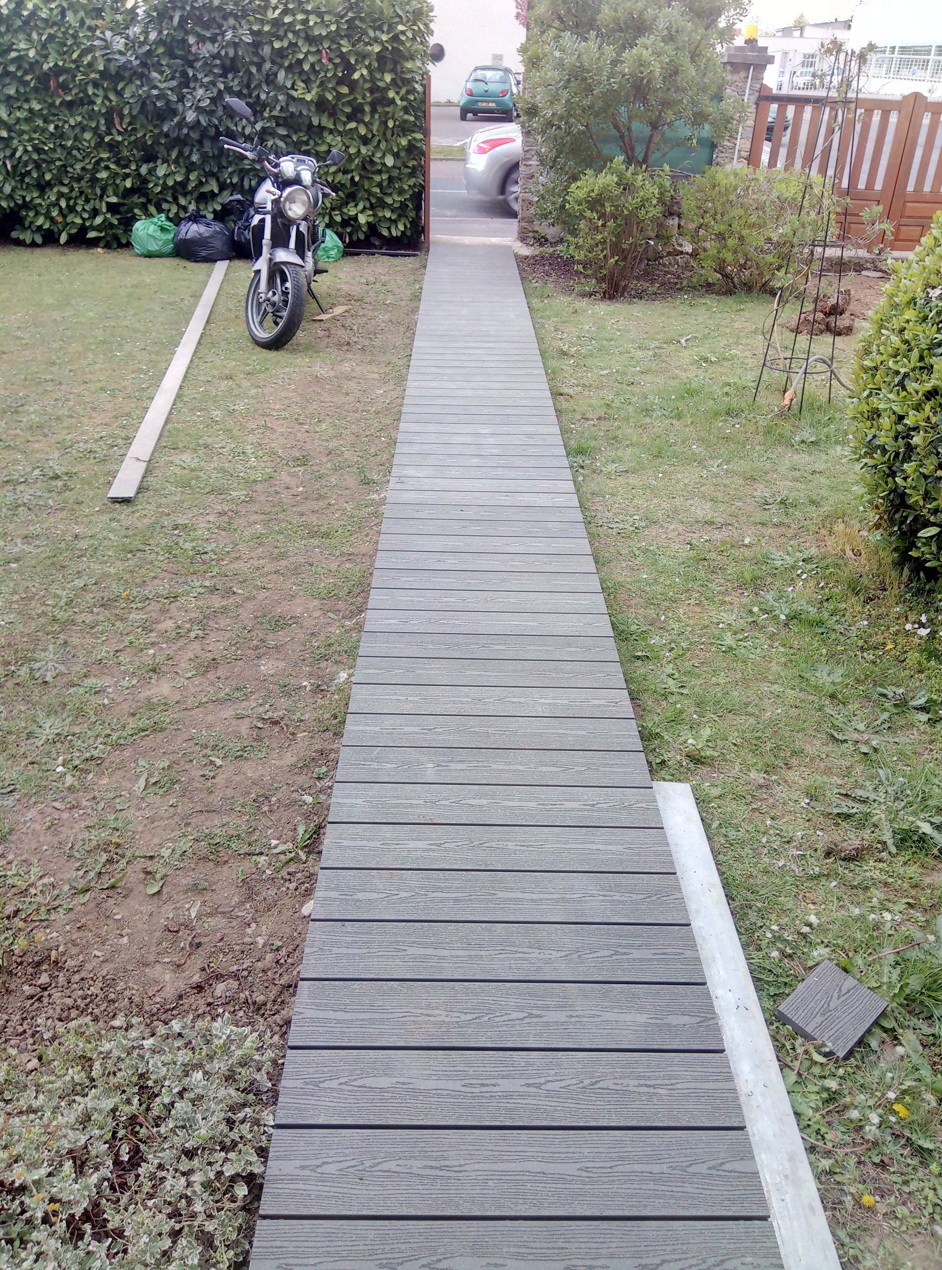 Une passerelle en bois traverse une cour herbeuse vers une allée avec un scooter garé à proximité.