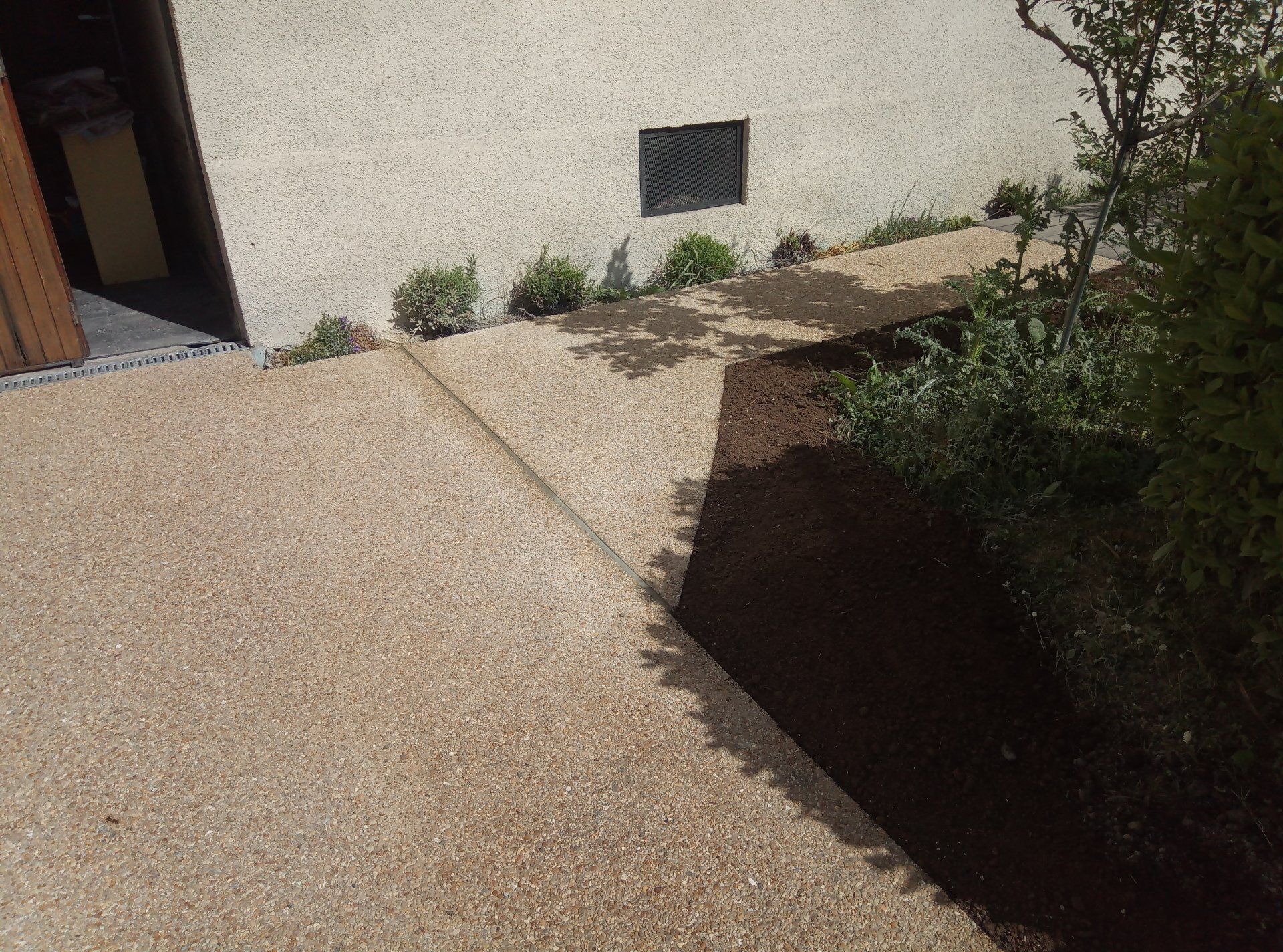 Chemin en béton moucheté beige à côté d'une bande de terre brun foncé avec des buissons verts et un bâtiment.