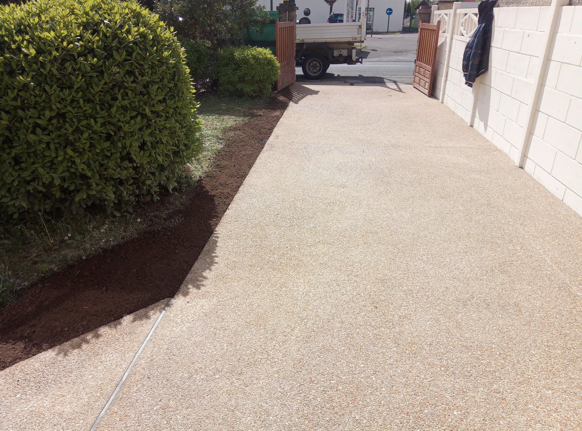Allée avec surface en gravier, un buisson vert et une bordure brune à gauche et un mur à droite.