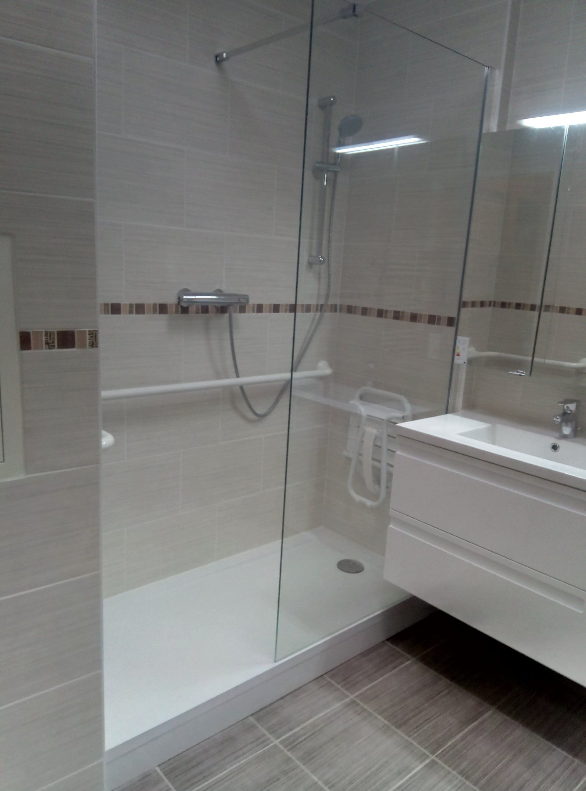 Salle de bain accessible : douche avec cabine de verre, barre d'appui et vanité blanche avec lavabo.