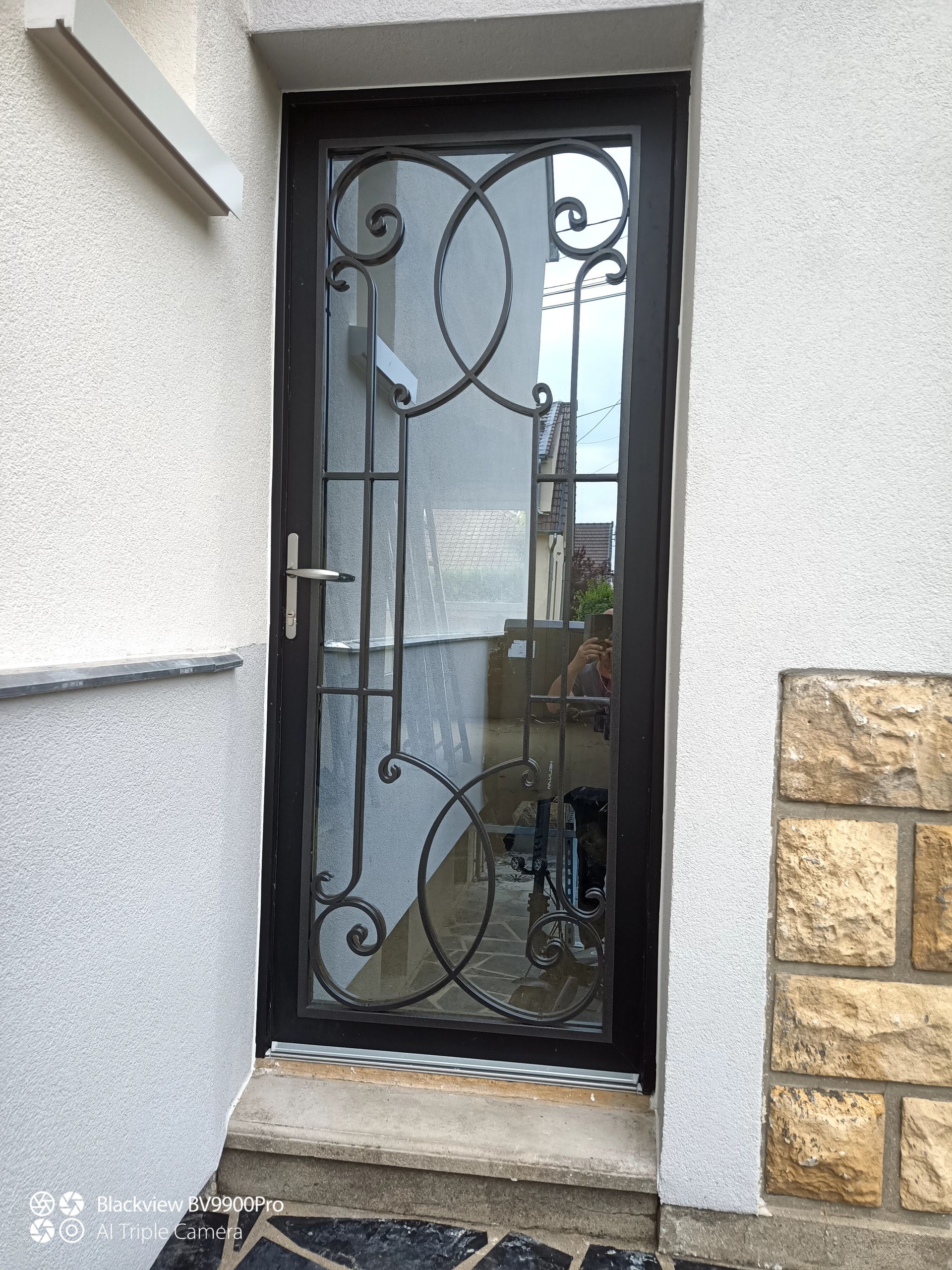 Porte d'entrée en métal noir et verre avec ferronnerie décorative, insérée dans un extérieur blanc et pierre.