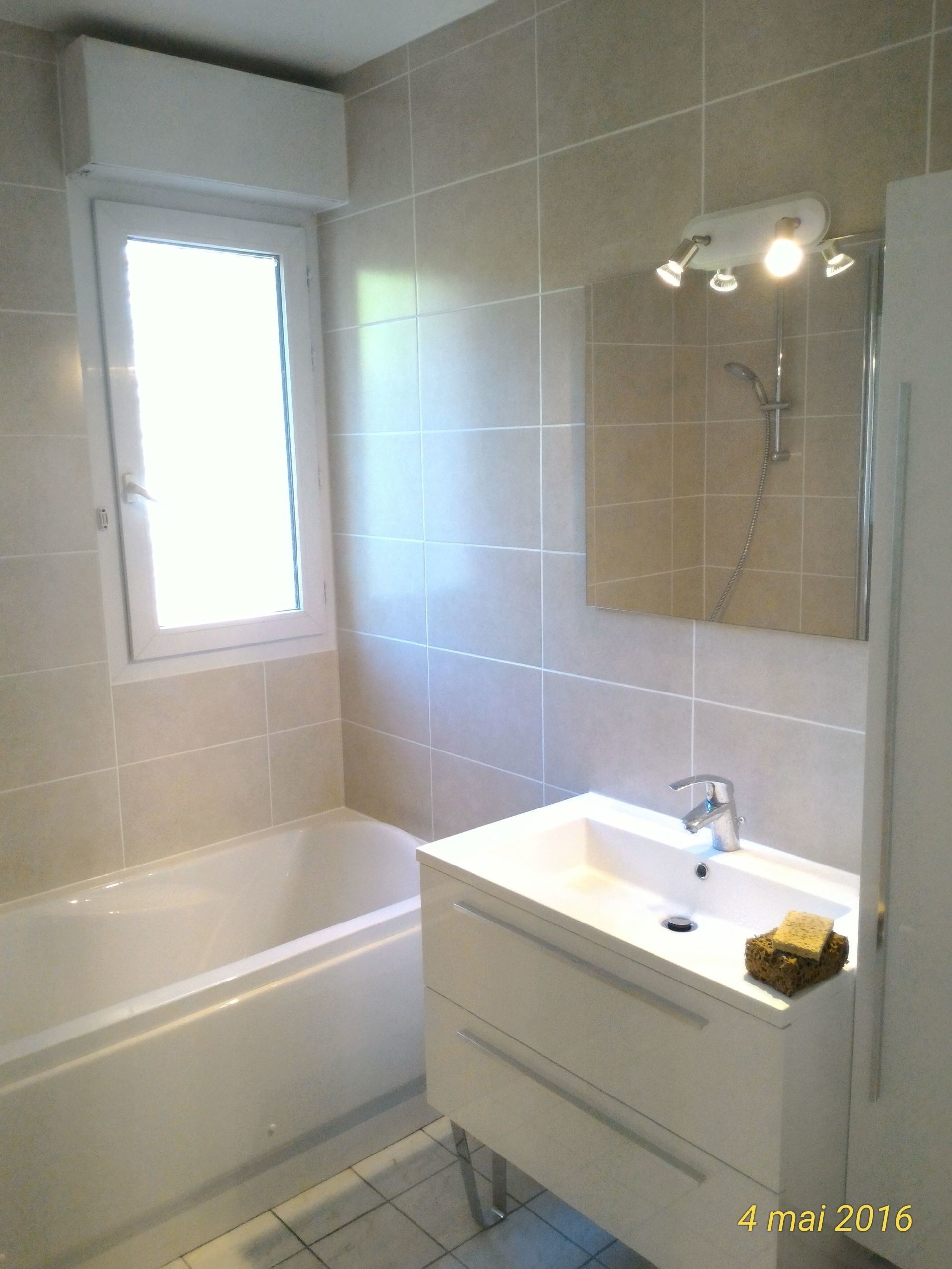 Salle de bain avec lavabo blanc, baignoire et murs carrelés beiges ; la lumière naturelle pénètre par une fenêtre.