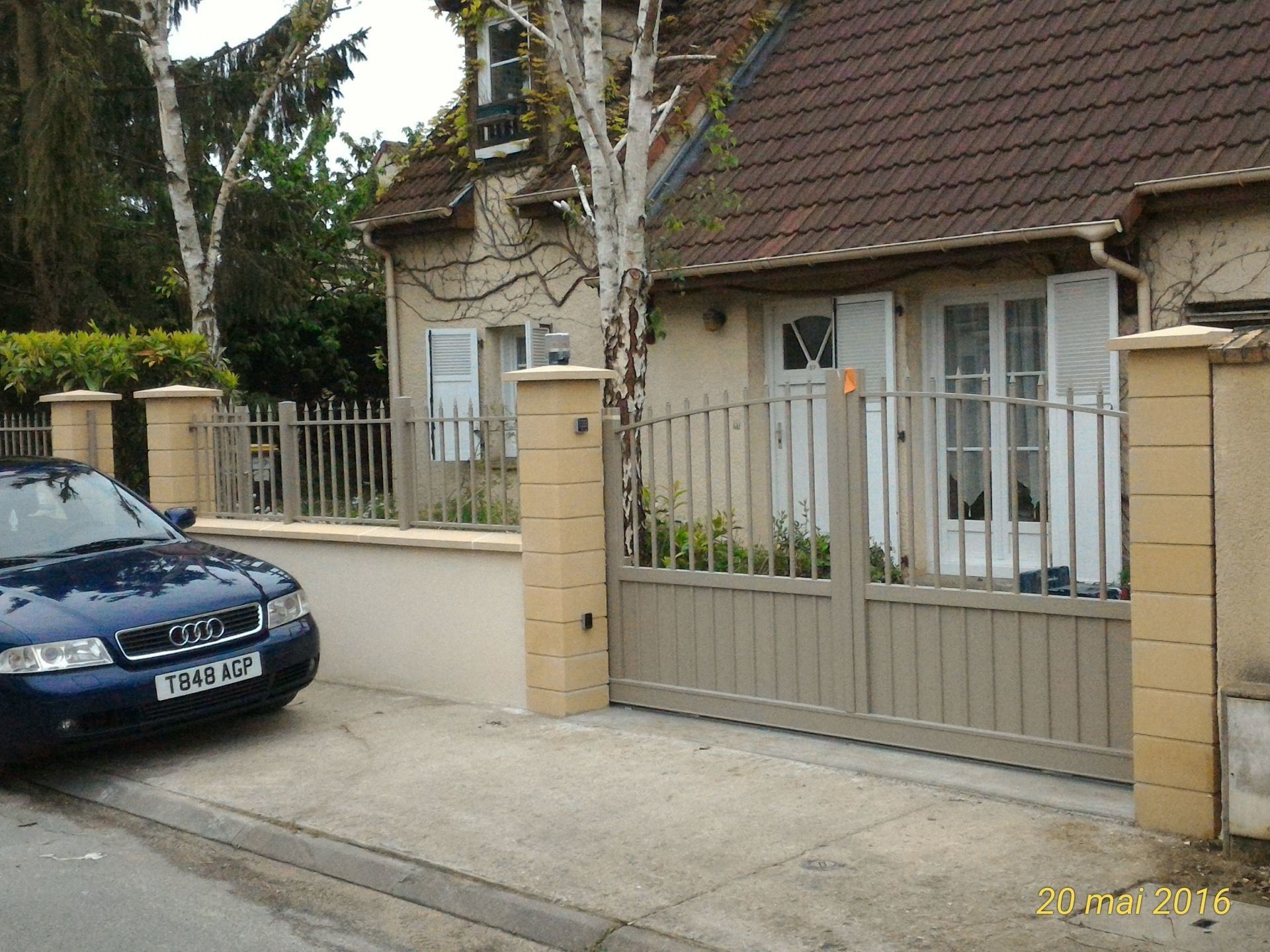 Une voiture bleue garée devant une maison beige avec une allée fermée et une clôture de couleur claire.