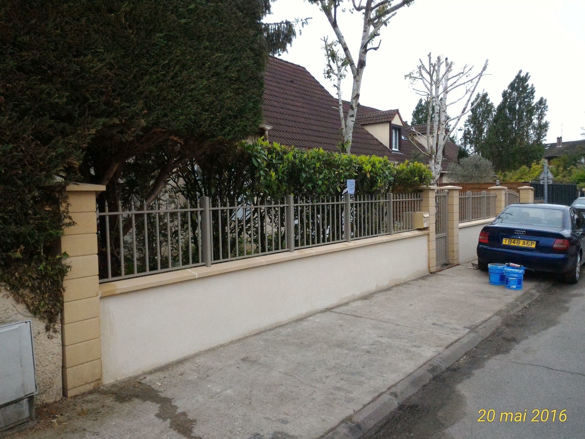 Un mur beige avec une clôture métallique devant une haie verte, rue et voiture à droite.