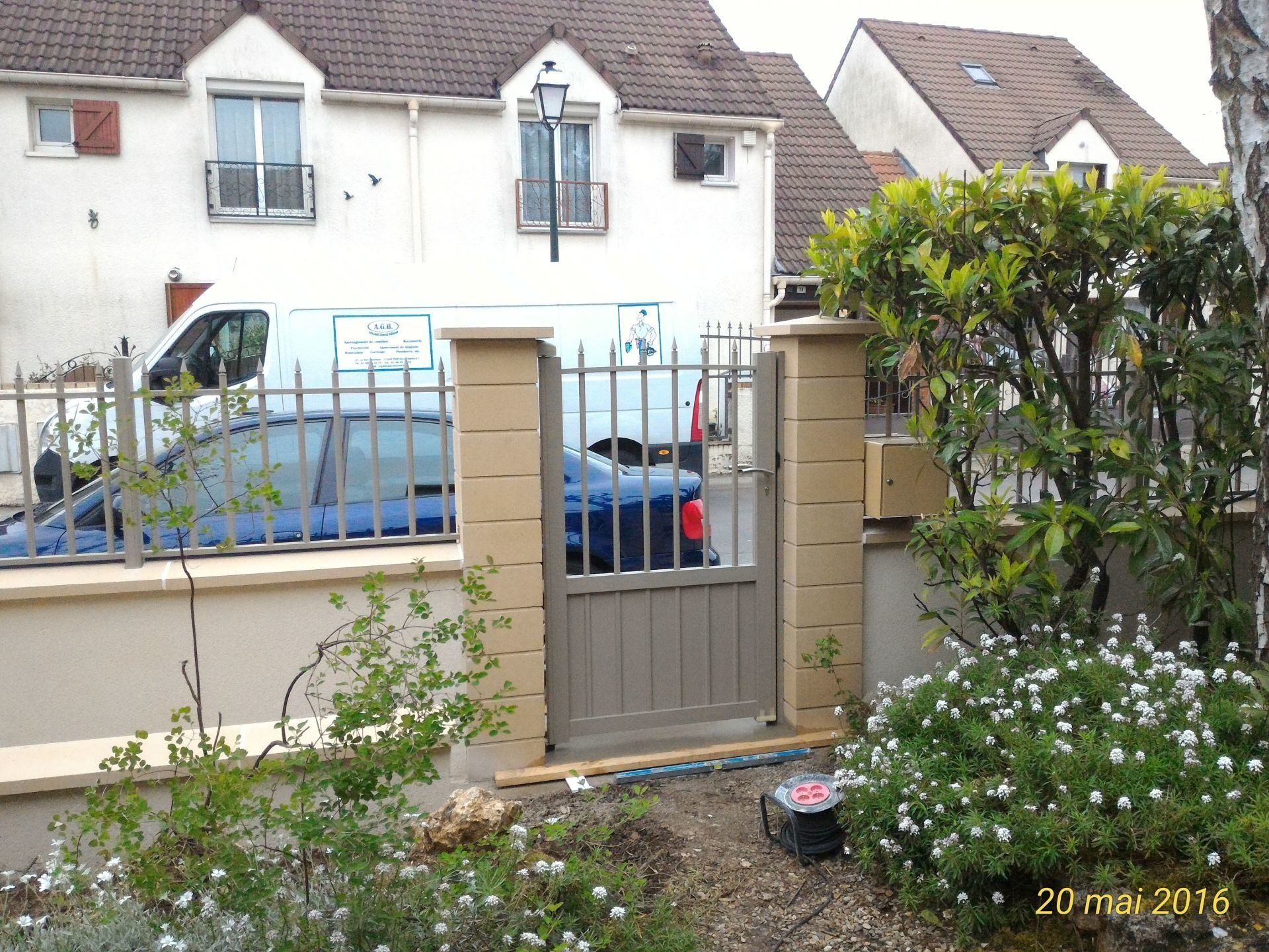 Une entrée fermée avec des piliers en pierre beige, une clôture en métal et une vue sur les maisons en arrière-plan.