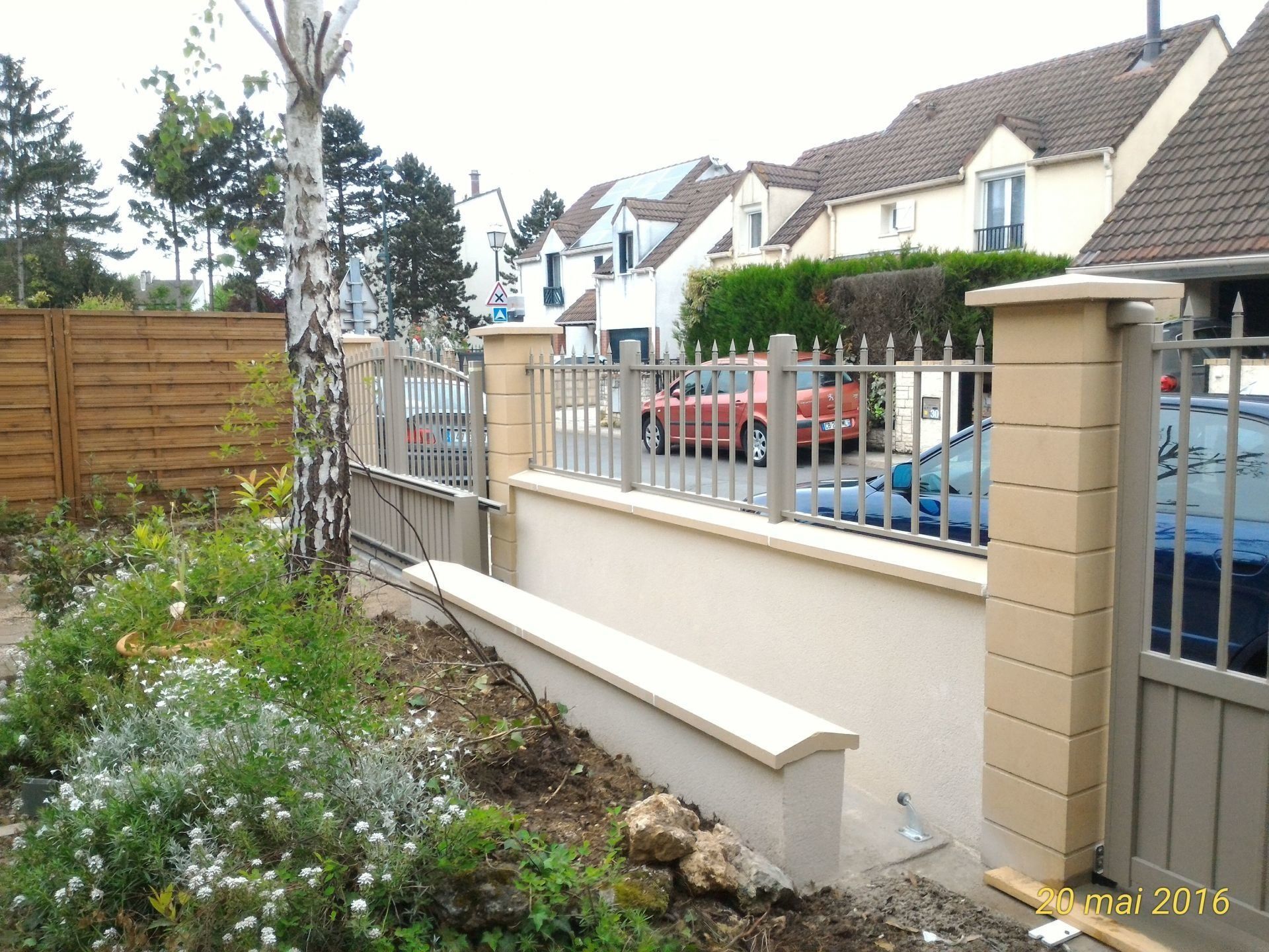Une scène de rue résidentielle avec une clôture de couleur beige, un muret et un portail devant les maisons.