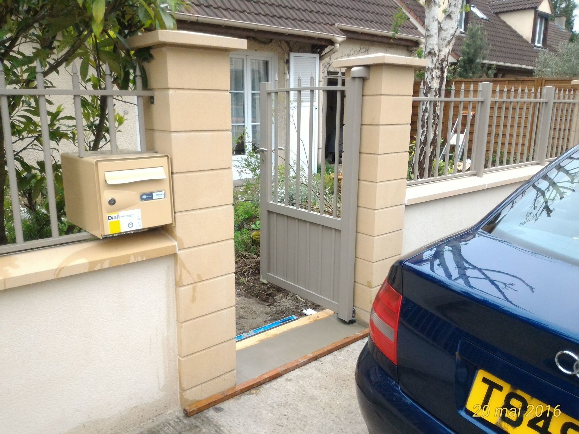 Portail d'entrée avec piliers beiges, boîte aux lettres et voiture bleue garée devant.