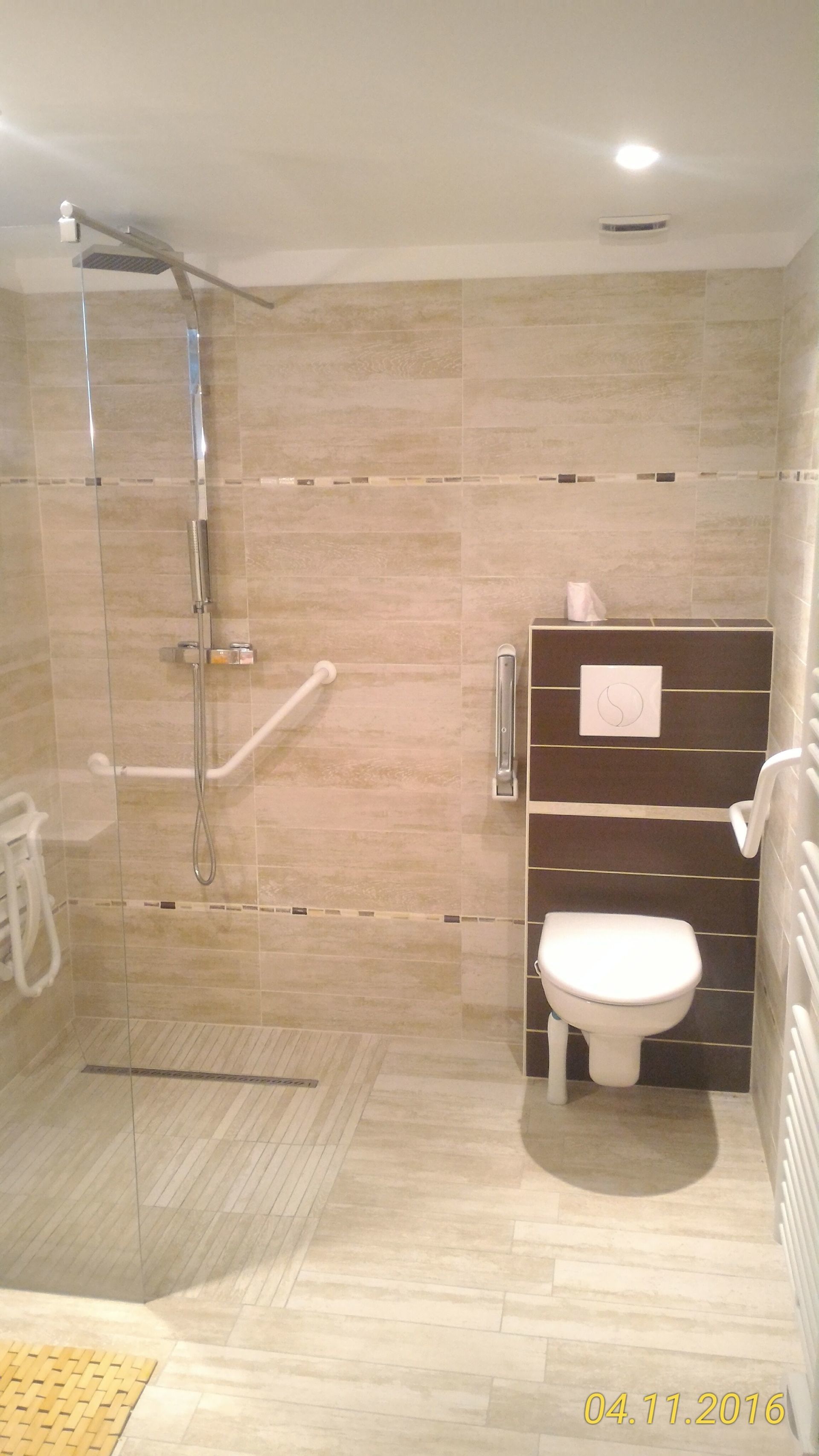Salle de bain accessible aux personnes à mobilité réduite avec douche, WC et barres d'appui, tons beige et marron.