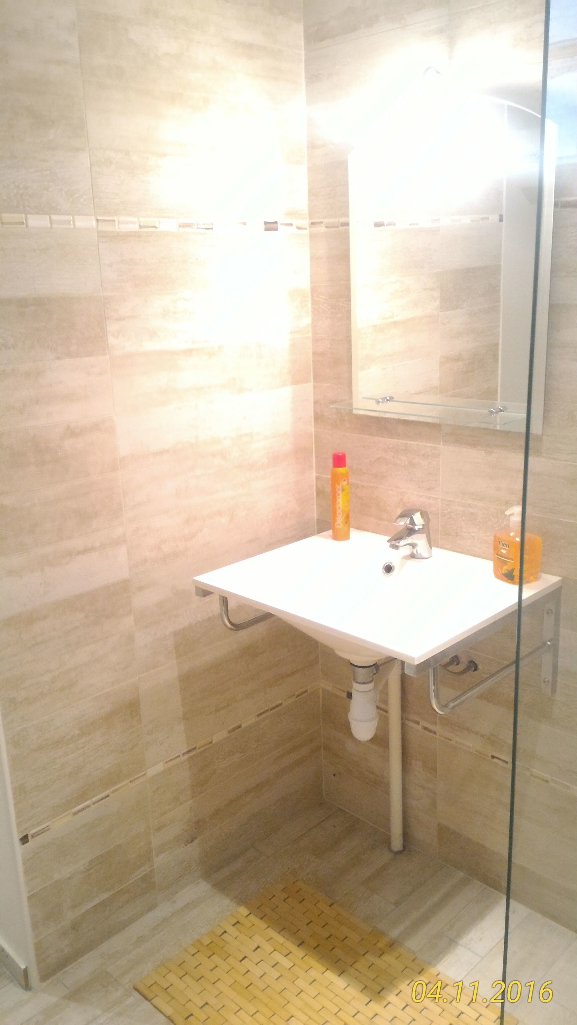 Salle de bain avec un lavabo mural blanc, un miroir et des murs beiges texturés, un tapis jaune et deux articles de toilette.