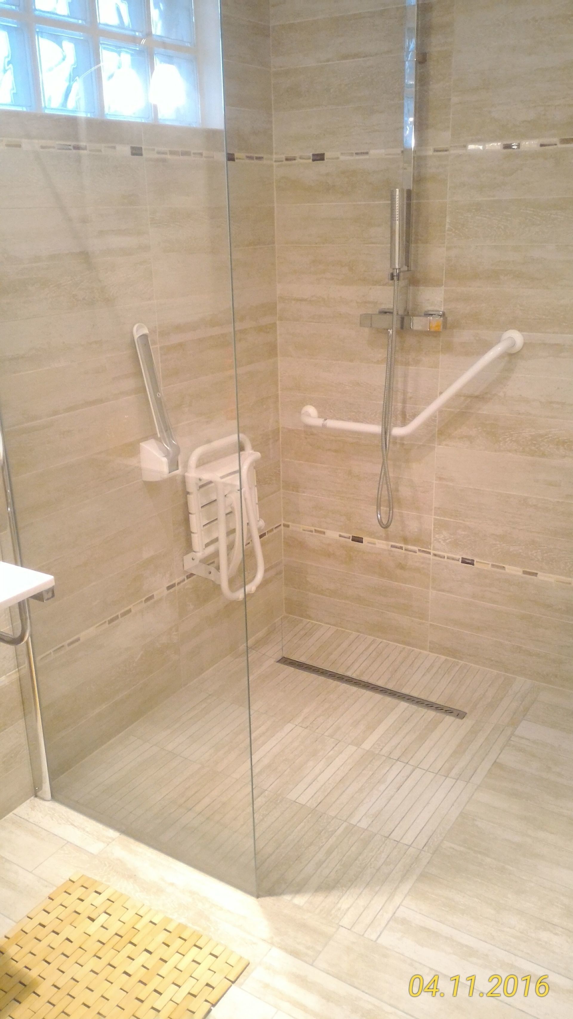 Douche accessible avec barres d'appui et siège rabattable. Carrelage beige, paroi en verre transparent.