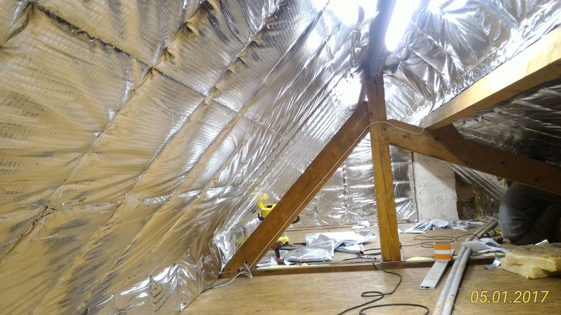 Intérieur du grenier avec isolation en feuille, poutres en bois et outils, partiellement éclairé par la lumière du jour.