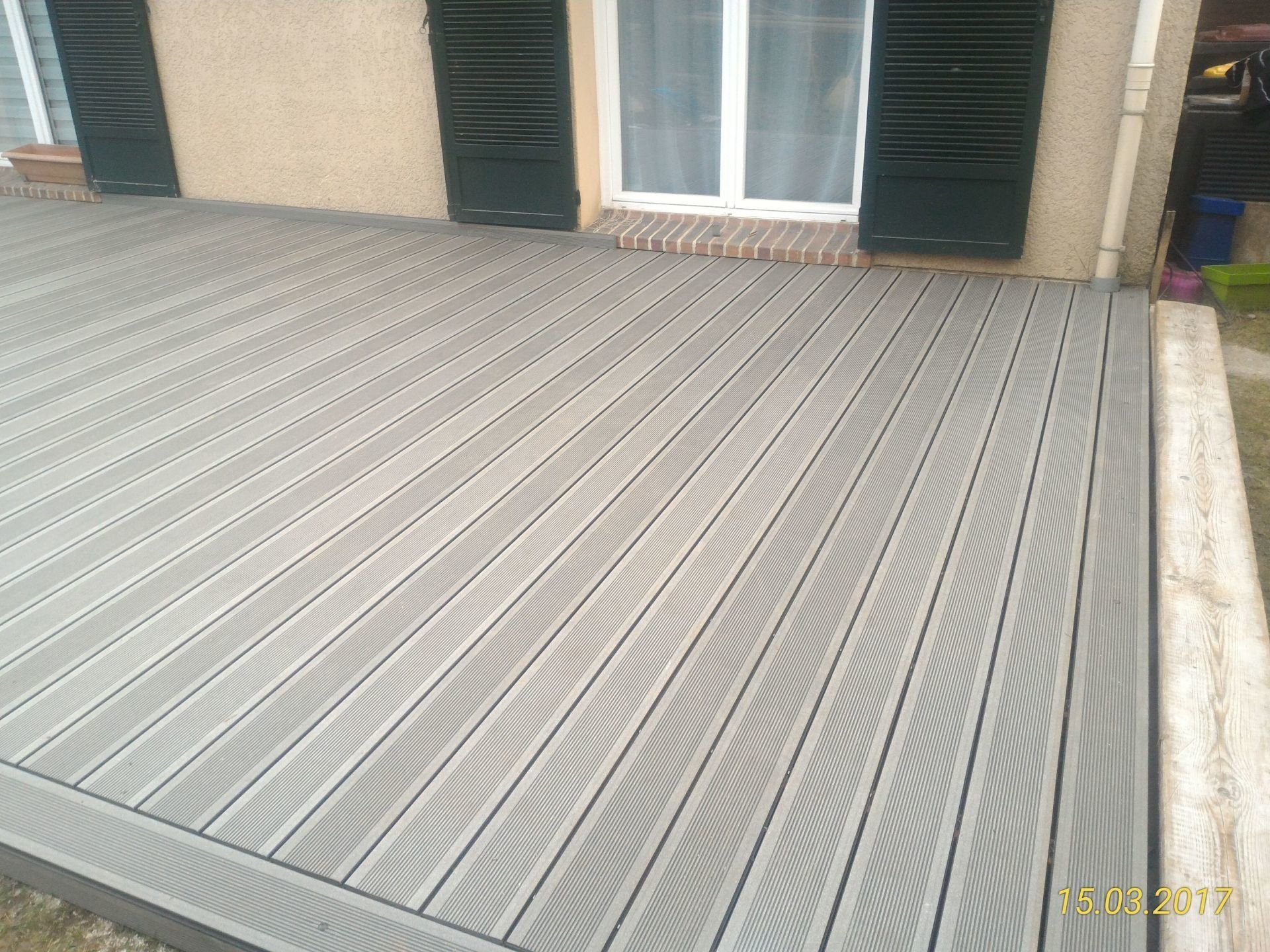 Terrasse composite grise devant une maison avec une fenêtre et des volets fermés.