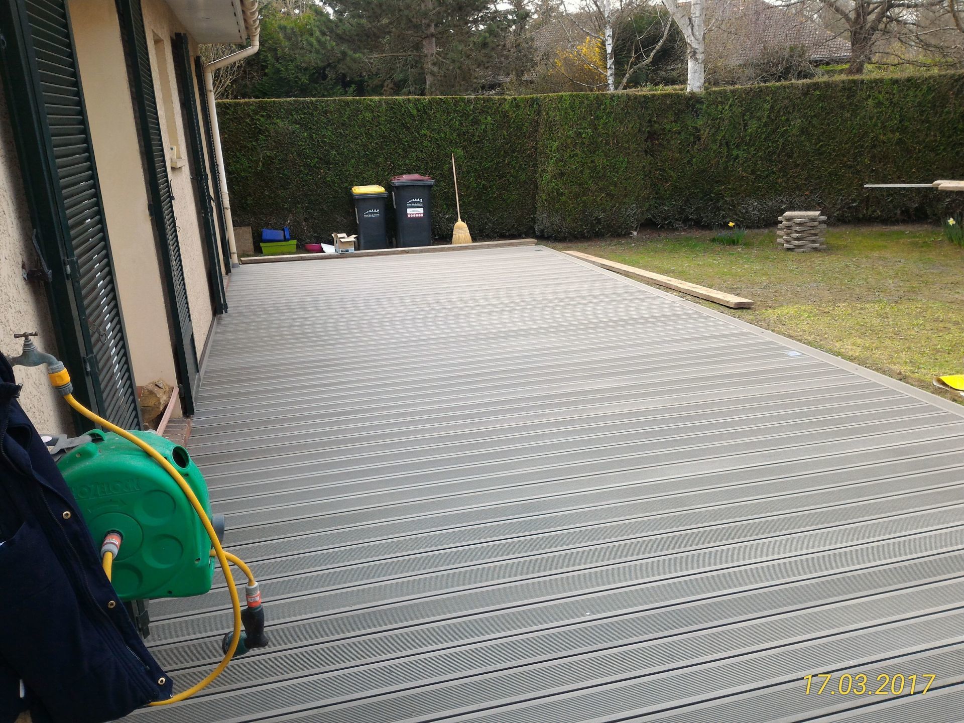 Patio à motifs gris à côté d'une maison, avec réservoir vert, poubelles et pelouse.