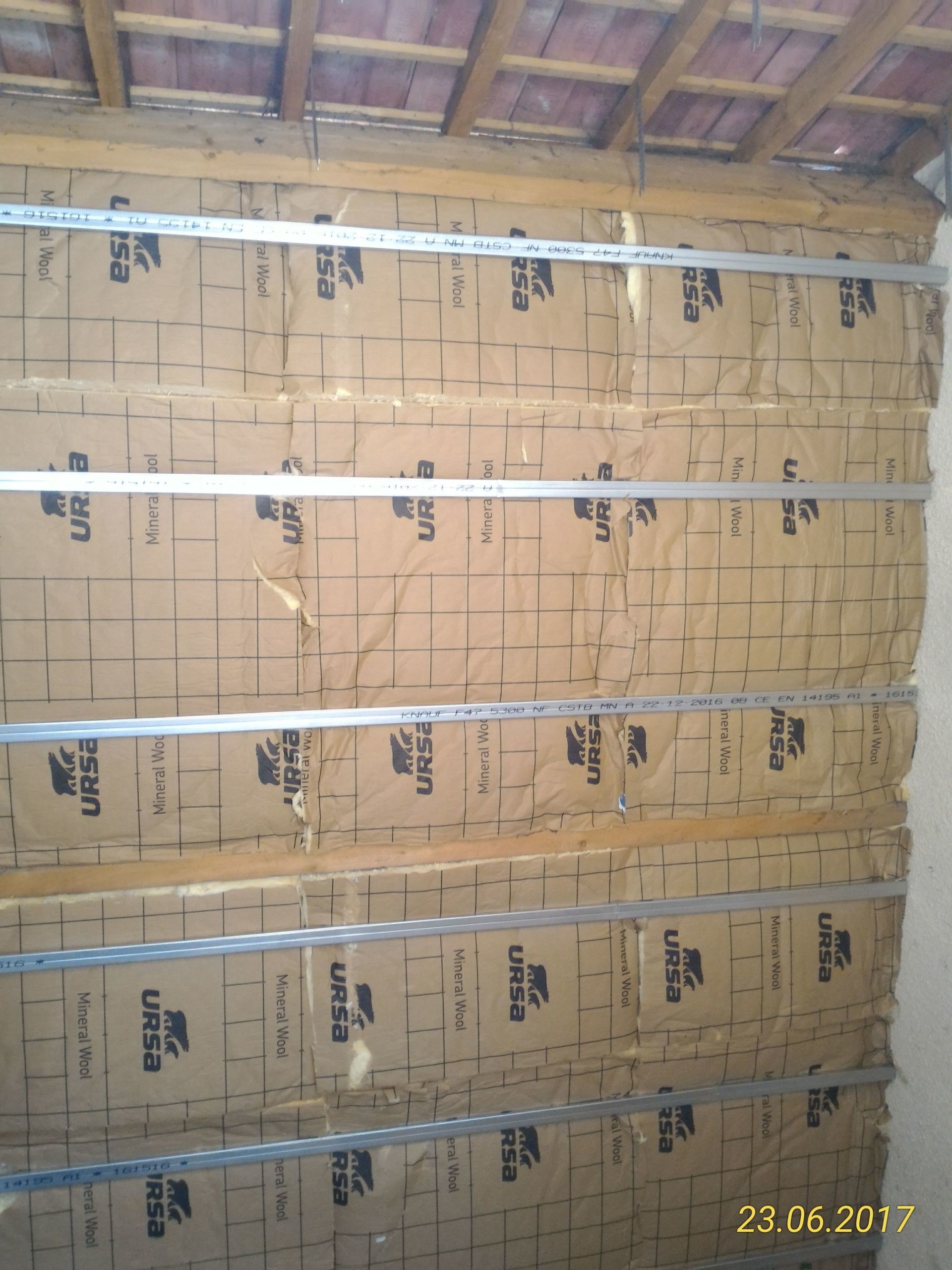 Isolation murale recouverte de papier kraft, soutenue par des rails métalliques, sous une toiture en bois et tuiles.