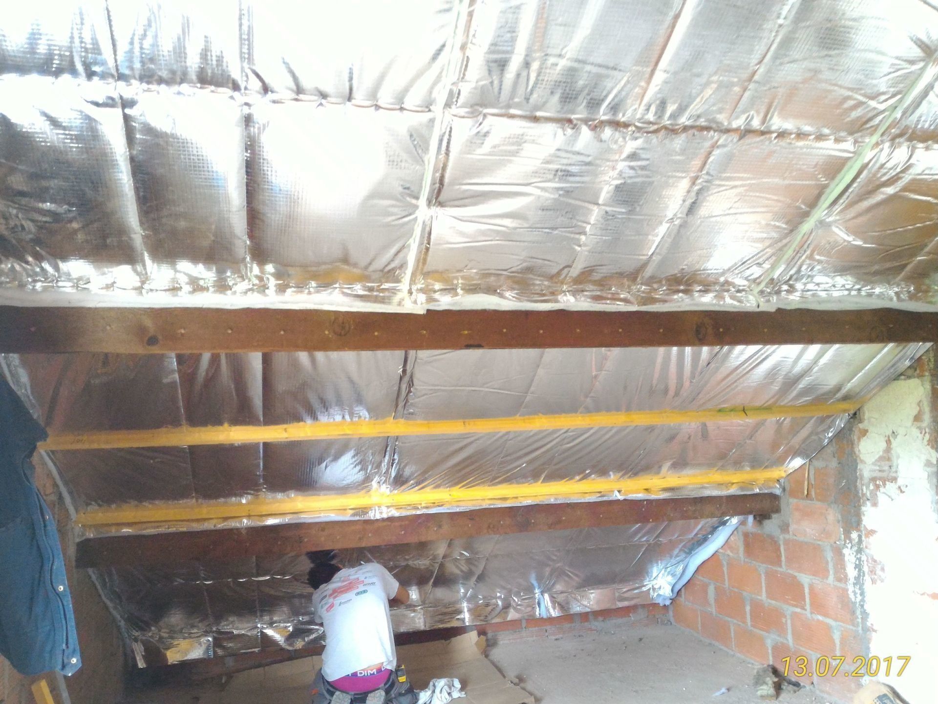 Personne installant une isolation réfléchissante sur un plafond en pente avec des poutres en bois et des murs en briques.