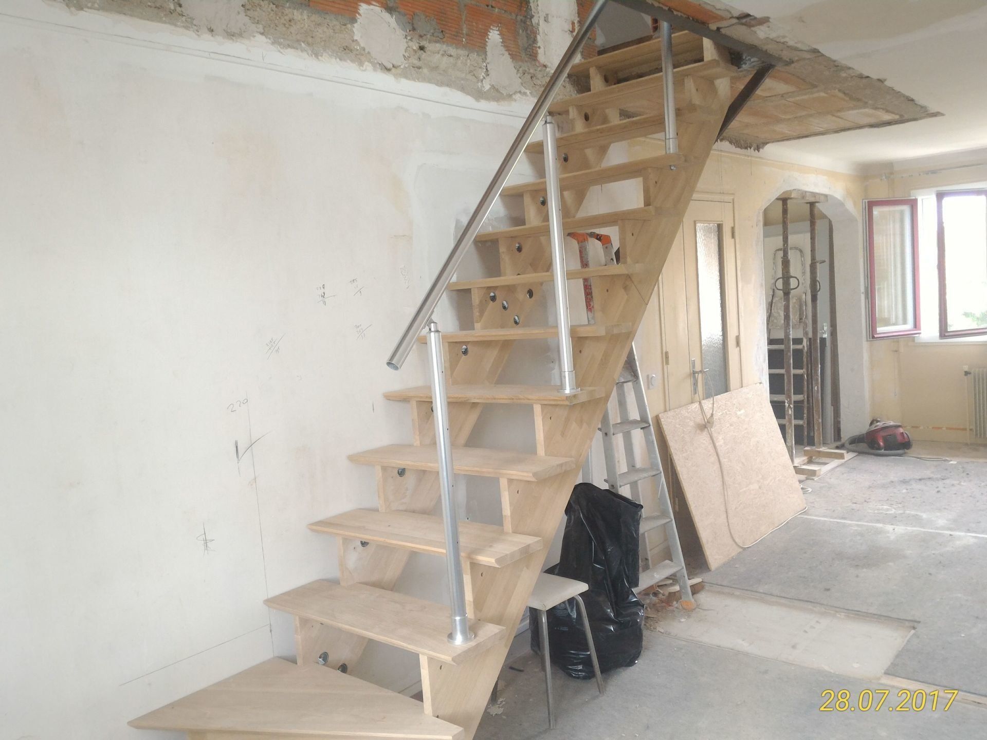 Escalier en bois en construction avec rampe en métal, dans une pièce aux murs et au sol non finis.