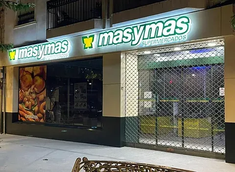 Fotografía nocturna de un supermercado Masymas con señalización luminosa. La entrada está parcialmente cerrada, mostrando una puerta de seguridad metálica.