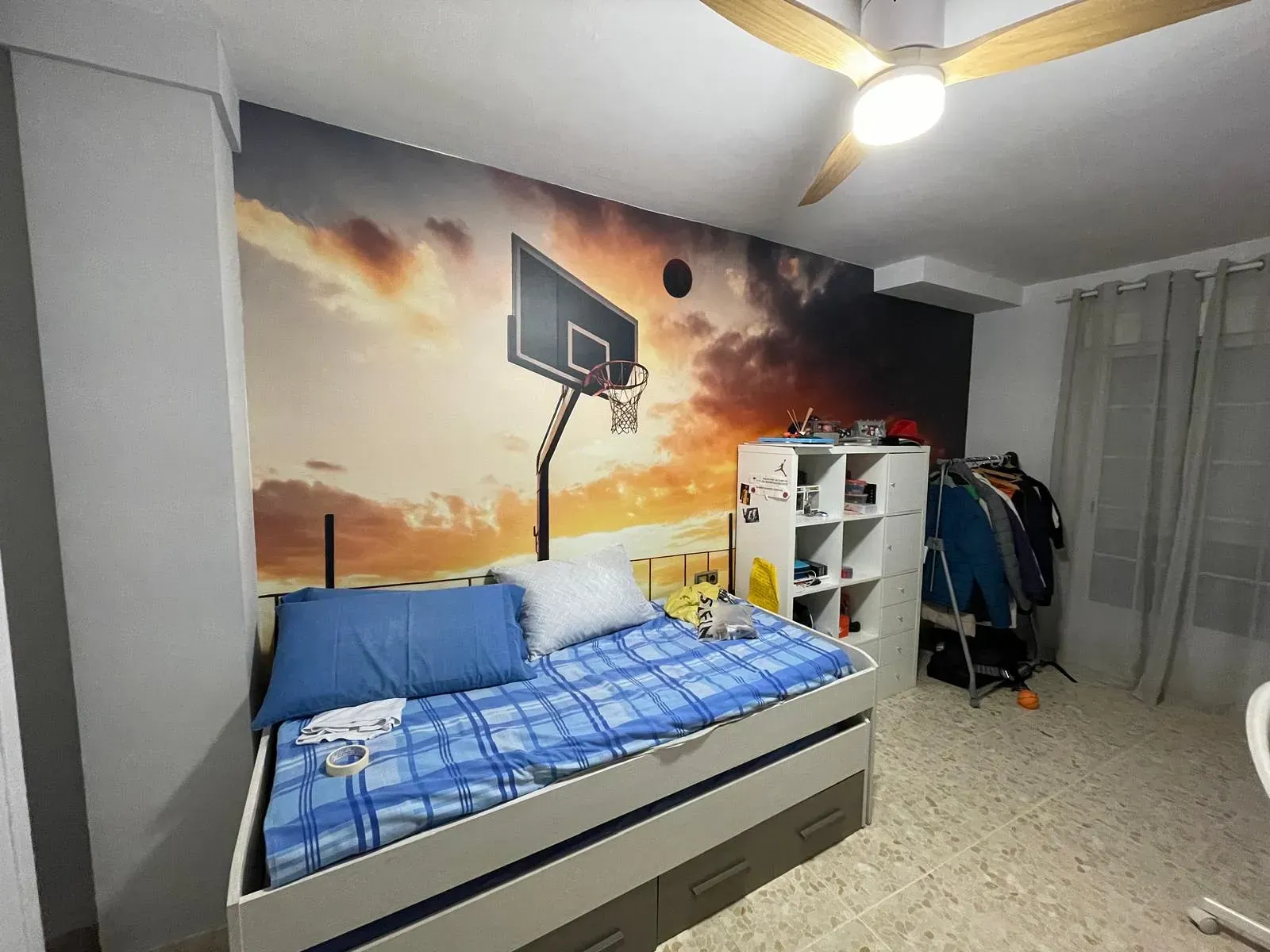 Dormitorio con mural de baloncesto. Cama con ropa de cama azul, cómoda blanca y perchero.