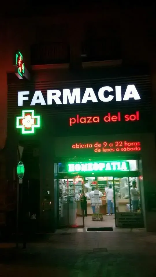 Una farmacia de noche, con el letrero "Farmacia Plaza del Sol" y una cruz verde. La fachada está iluminada y se ve "HOMEOPATIA" en el interior.