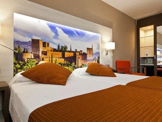 Camas individuales en una habitación de hotel con cabecero decorado con un mural de la Alhambra. Se ven ropa de cama naranja, lámparas y una mesita de noche.