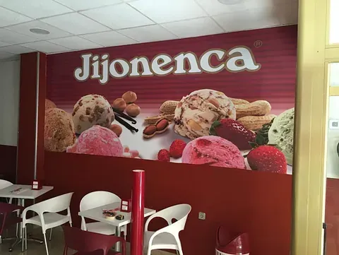 Interior de una heladería jijonenca. Un gran cartel muestra el logotipo y varios sabores de helado. Mesas y sillas instaladas.