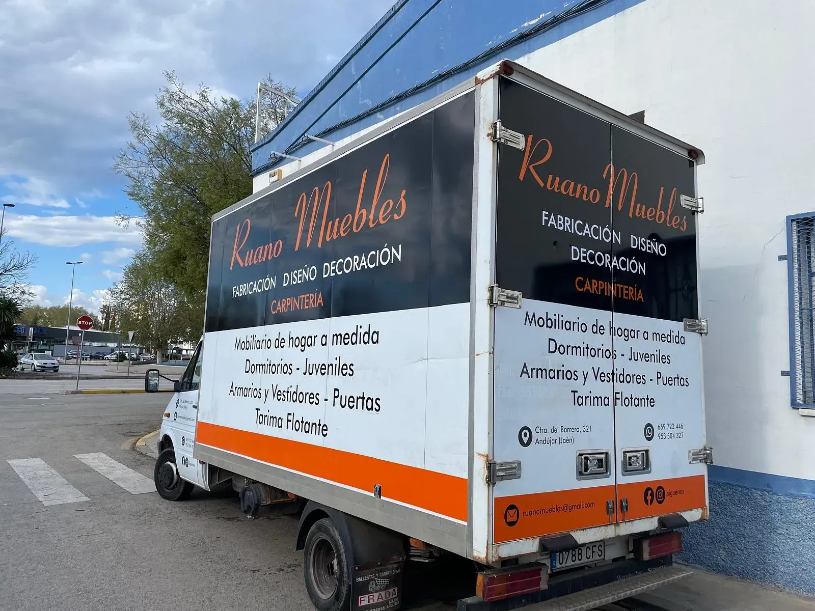 Un camión de reparto blanco estacionado en una esquina con la marca "Ruano Muebles". Presenta texto en negro, naranja y blanco que promociona sus servicios de muebles.